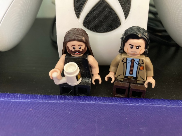 Tho lego figurines of people. On the right is Jonathan Van Ness who has a bead and long hair. They are holding a lego mug. Beside JVN is a Loki lego figurine. He has shoulder length hair and is wearing close from the Loki show.